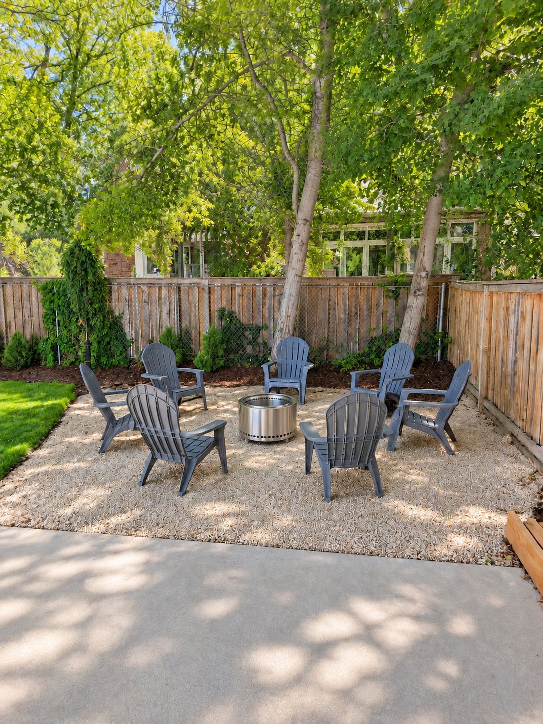 An outdoor seating area with six Adirondack chairs arranged around a metal fire pit, set on a gravel surface with a wooden fence in the background.