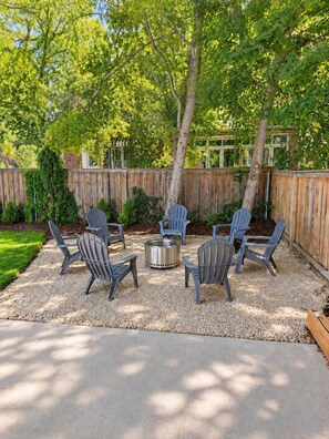 An outdoor seating area with six Adirondack chairs arranged around a metal fire pit, set on a gravel surface with a wooden fence in the background. - Newly Renovated Riverwoods Retreat — 4BR, 3BA (Orem)