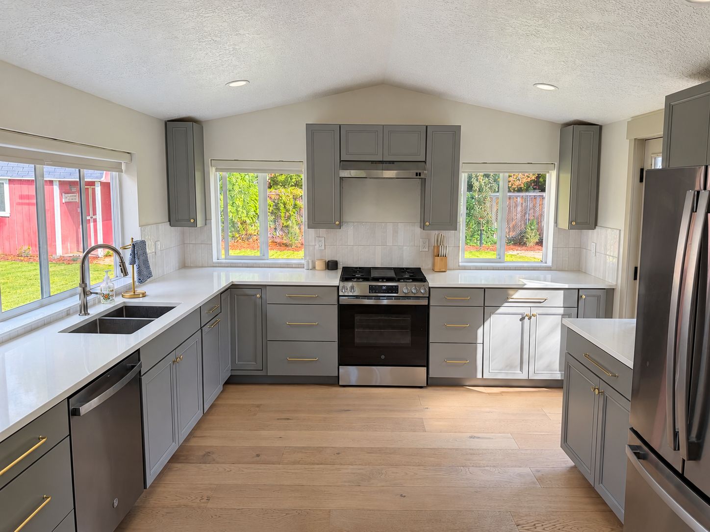 A modern kitchen with grey cabinets, stainless steel appliances, and a large window providing a view of the outdoors.