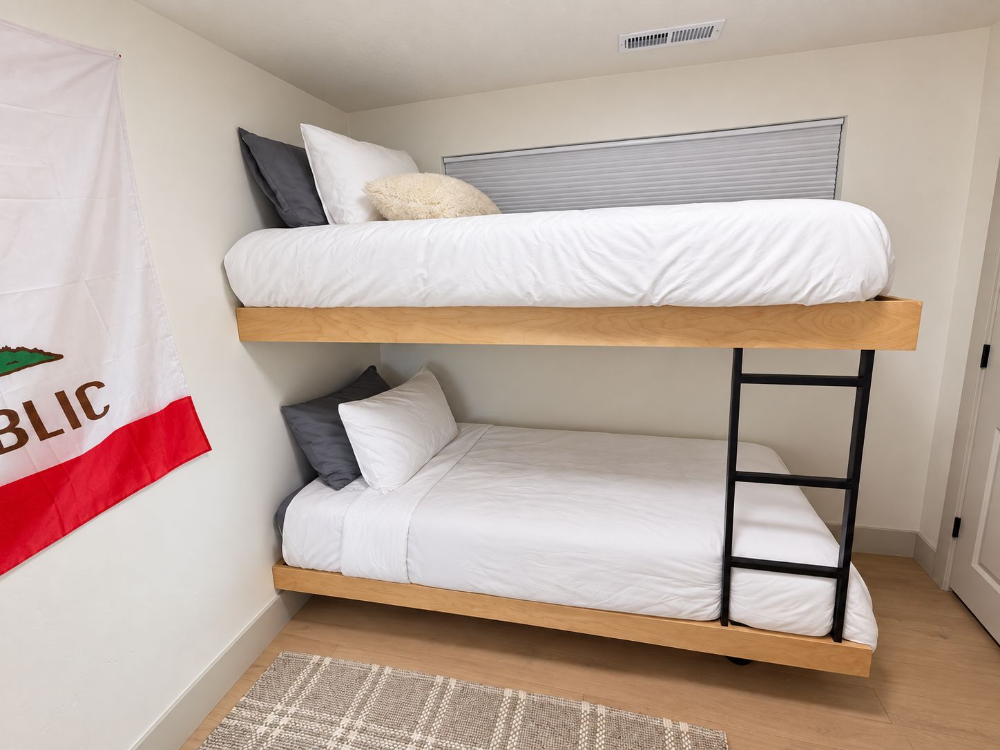 Ein Stockbett mit Holzrahmen und weißer Bettwäsche, ein kleiner Teppich auf dem Boden und eine an der Wand befestigte Flagge.