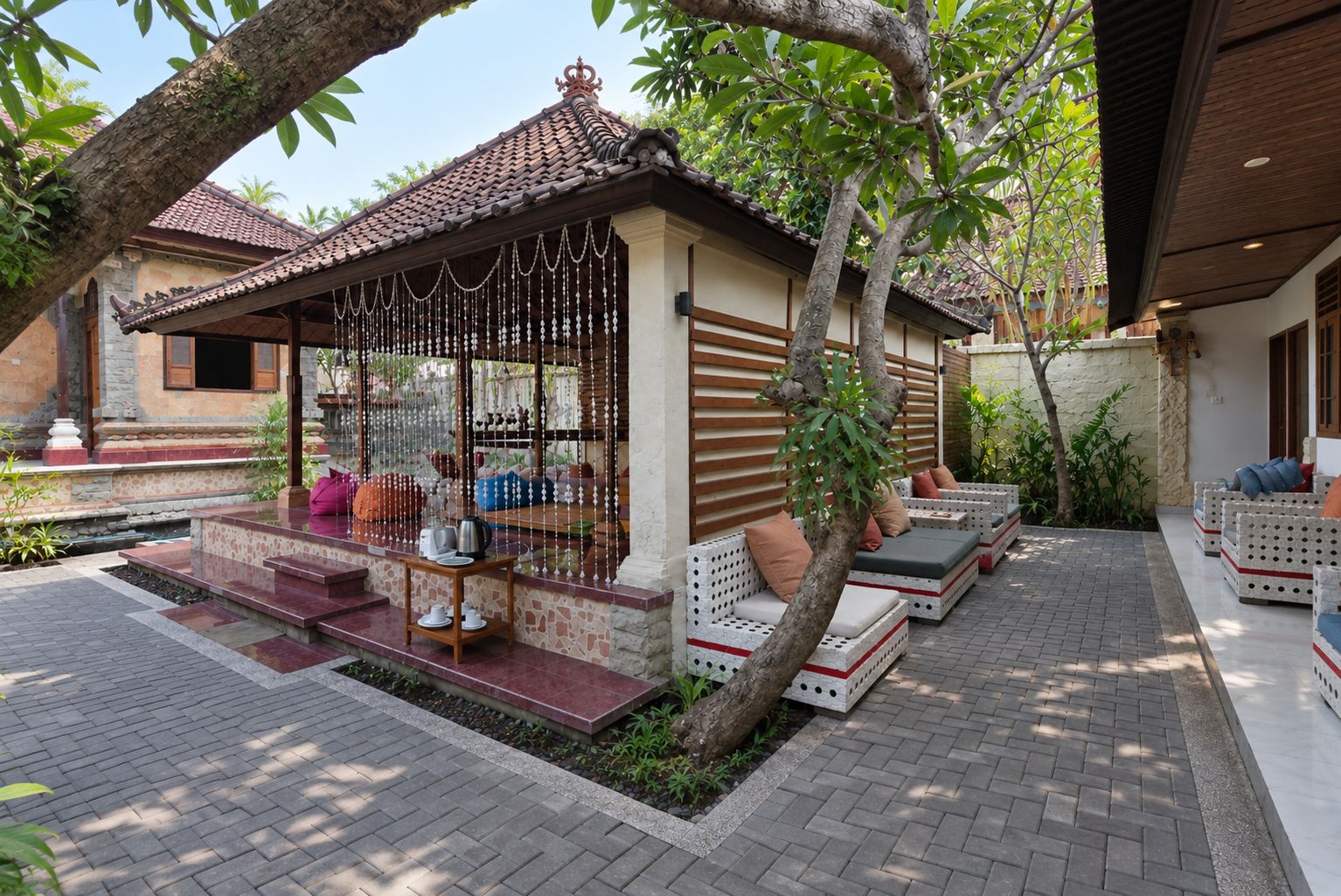 A covered outdoor seating area with a table and chairs, surrounded by plants and trees.