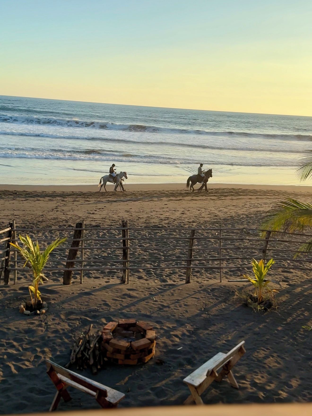 A beach with two people riding horses, a wooden fence, and a fire pit.