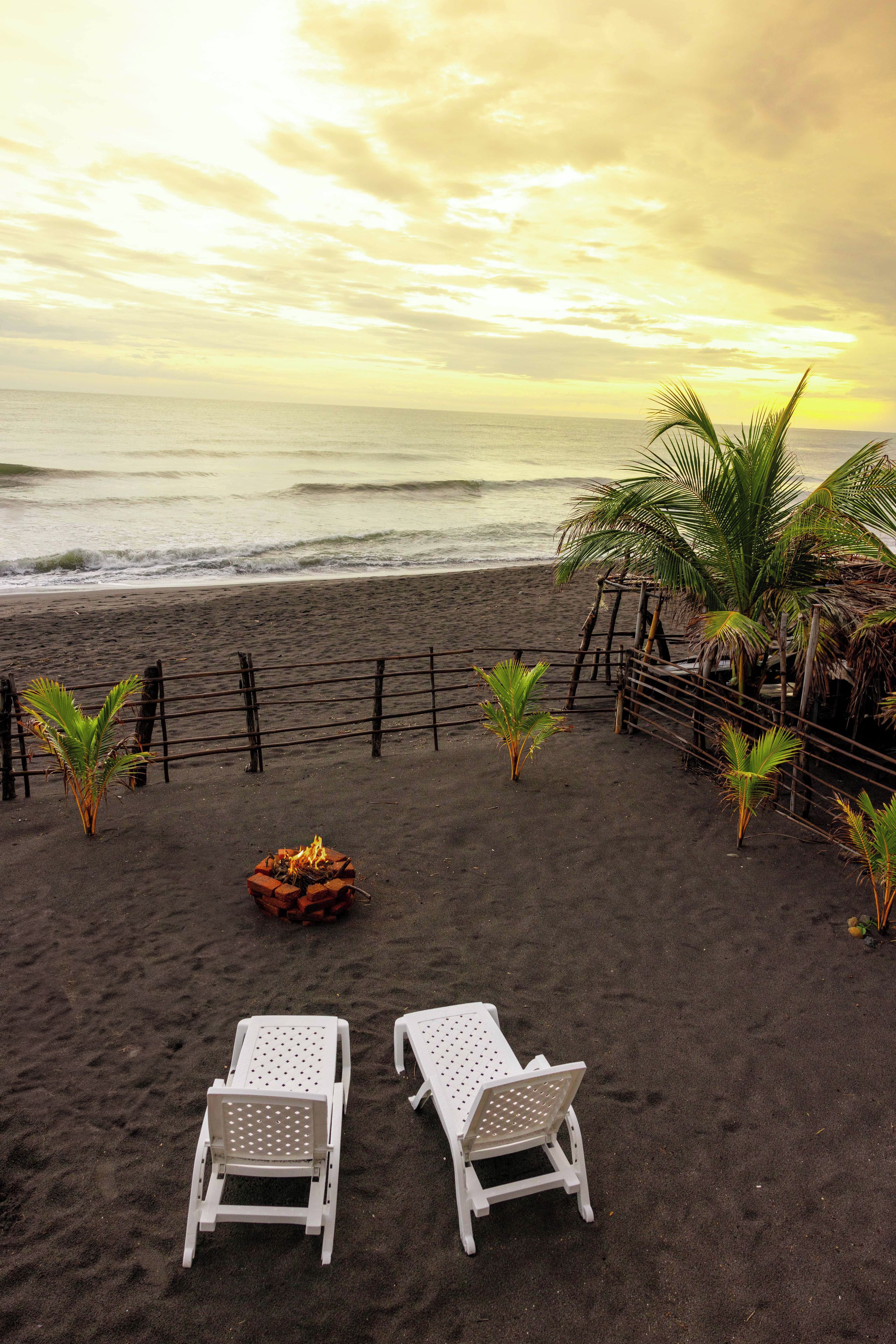 A beach with two white lounge chairs, a small fire pit, and a metal fence.