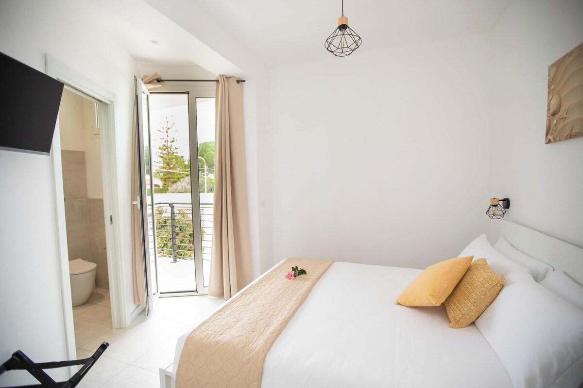 A bedroom with a bed, a bedside table, a hanging light fixture, and a view through a door.
