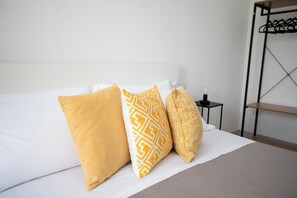 A bed with three pillows, a bedside table with a lamp, and a clothing rack. - Golden Bay Apartments (Arenella)