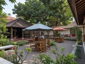 An outdoor seating area with wooden tables and chairs, covered by umbrellas, surrounded by greenery and a building with a tiled roof. - OYO 95446 Golden Homestay (Medan)