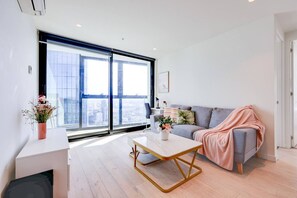 Uma sala de estar moderna com uma janela grande, um sofá cinzento, uma mesa de centro branca e uma mesa lateral com um vaso de flores.