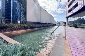 Um espelho de água com guarda-corrimão de metal, rodeado por vegetação e edifícios modernos.