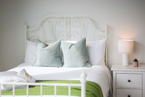 Cama bem arrumada com cabeceira de metal branca, dois travesseiros, manta verde e mesa de cabeceira com abajur e uma pequena planta.