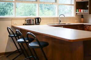 Een keuken met een groot schiereiland, barstoelen, een waterkoker en een raam met uitzicht op groen.
