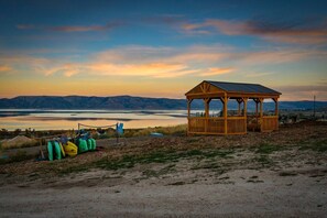 A wooden gazebo with a metal roof overlooking a lake, surrounded by a gravel area with green and yellow kayaks.