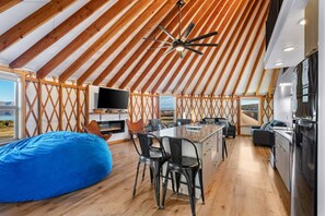A spacious interior with a high wooden ceiling, a central seating area with a fire feature, a kitchen area, and a large window offering a view.