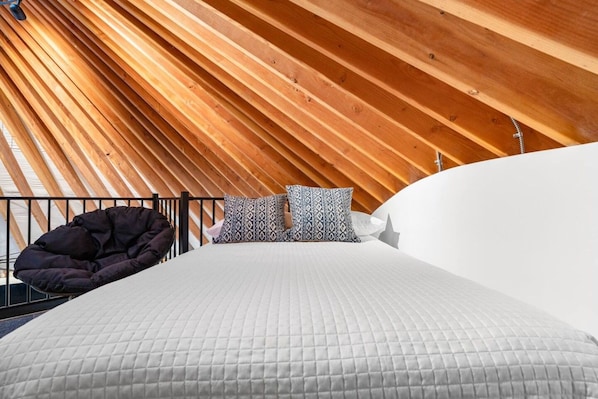 A room with a sloped ceiling, a bed with a quilted mattress, patterned pillows, and a black bean bag chair.