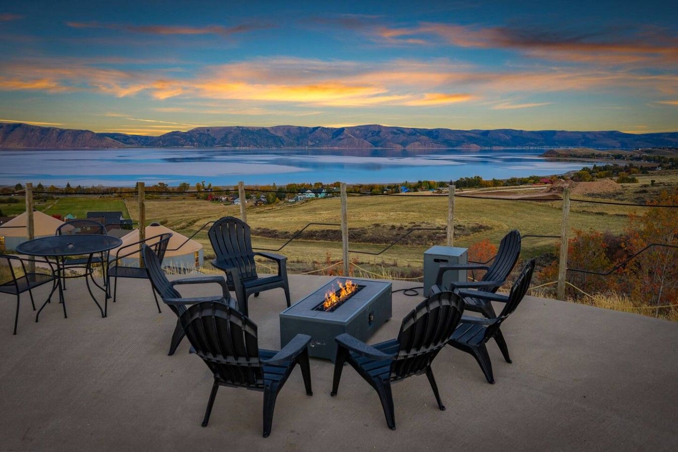 An outdoor patio with a fire pit, chairs, and a table overlooking a lake and mountains.