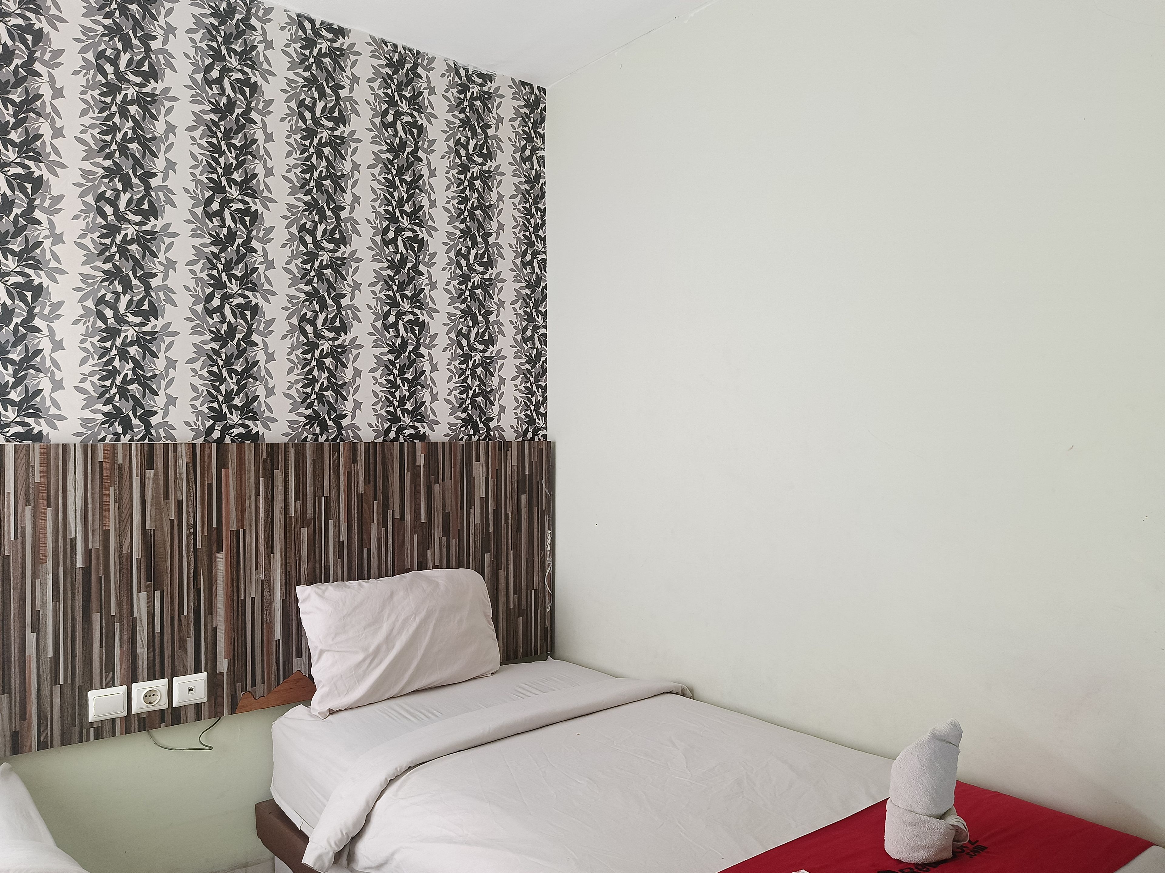A hotel room with a patterned wallpaper, a wooden headboard, a bed with white bedding, and a red accent pillow.