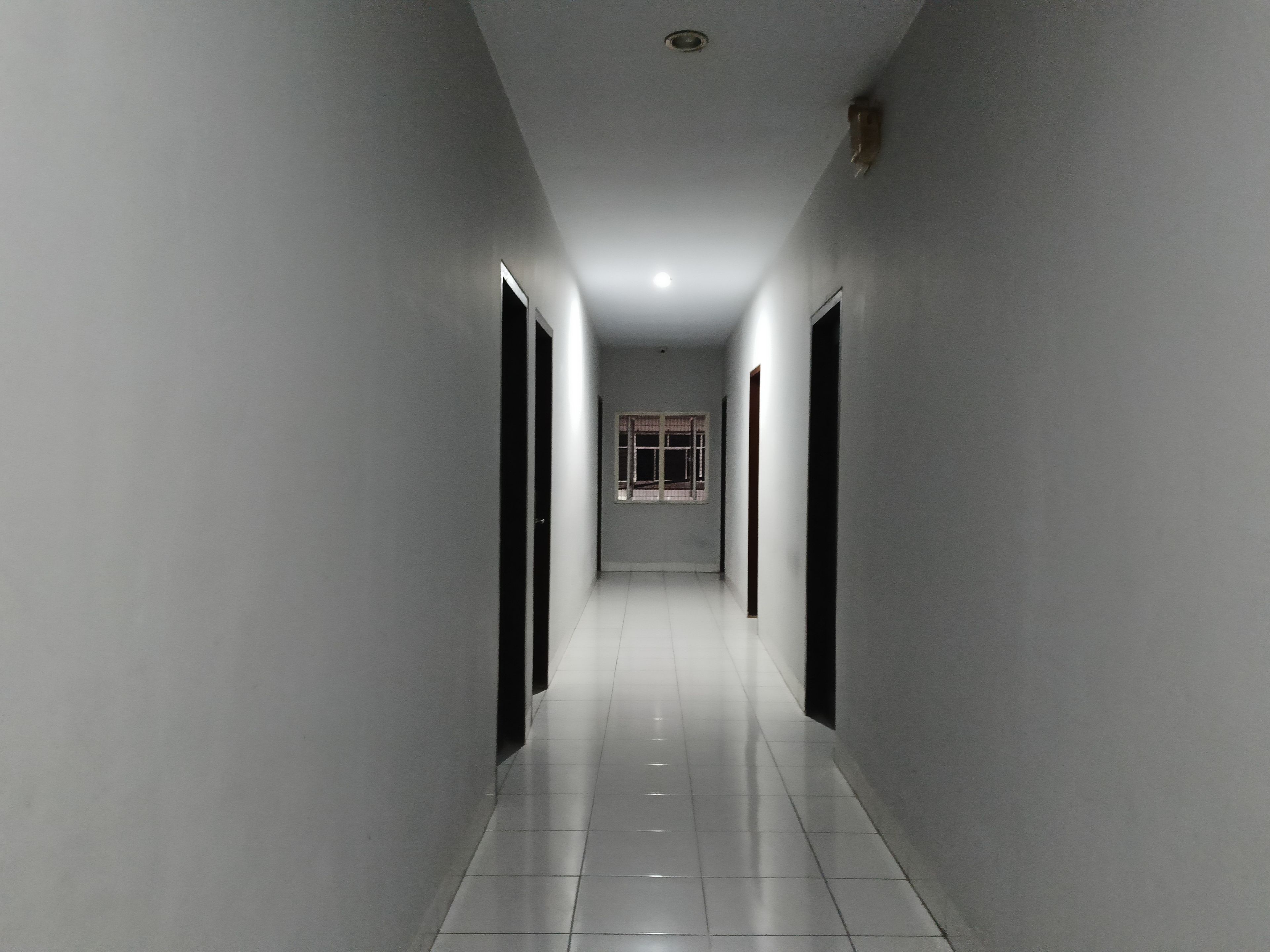 A corridor with white walls and a tiled floor, leading to a room with a window and a door.