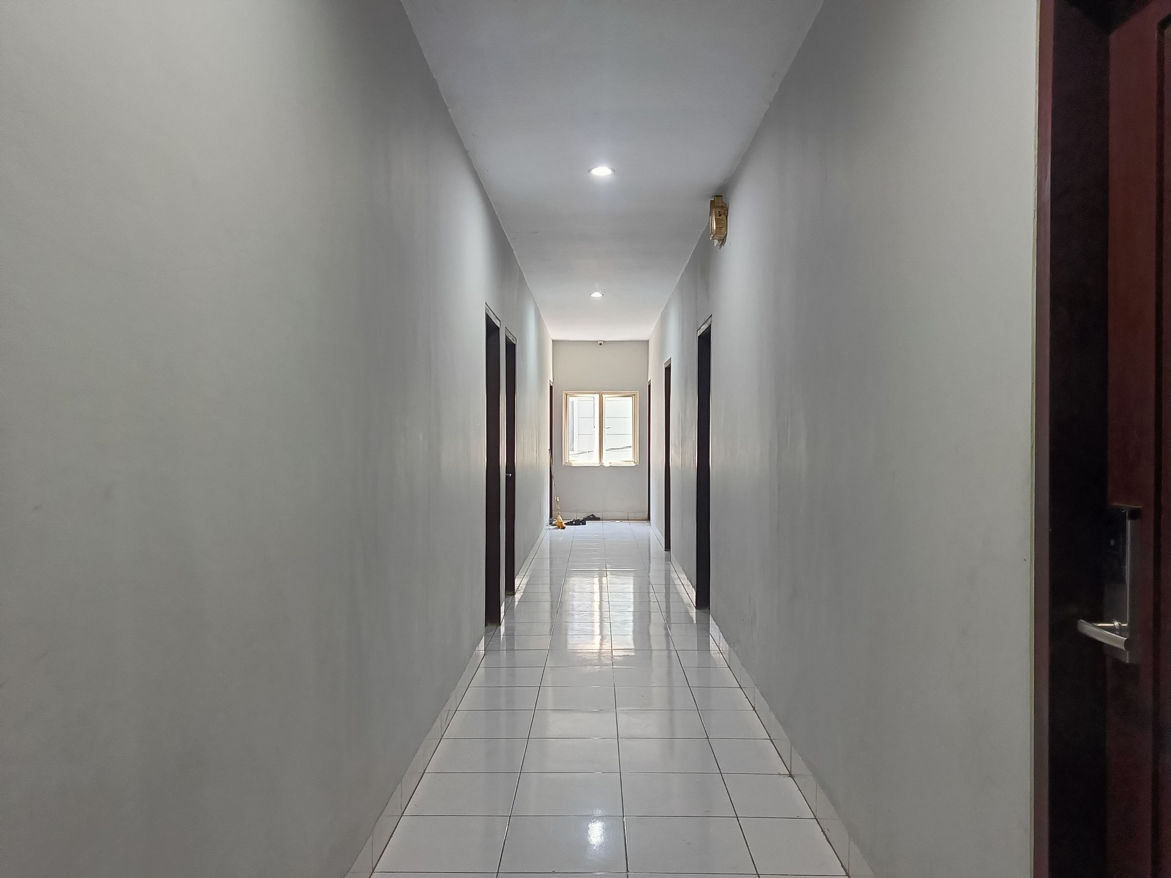A corridor with white tiled flooring, doors on both sides, and a window at the end.