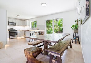 A modern kitchen with white cabinets, a large wooden table, and a view of lush greenery through the windows.