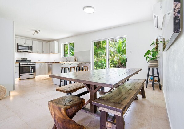 Una cocina moderna con armarios blancos, una mesa de madera grande y vistas a vegetación exuberante a través de las ventanas.