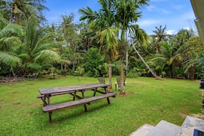 A grassy outdoor area with a wooden picnic table and chairs, surrounded by lush tropical vegetation and palm trees.