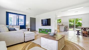 Een moderne woonkamer met een bank, salontafel en televisie. Een keuken met eettafel en stoelen is zichtbaar door een geopende deur.