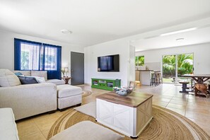 Ein modernes Wohnzimmer mit einer Couch, einem Couchtisch und einem Fernseher. Durch eine offene Tür ist eine Küchenzeile mit einem Esstisch und Stühlen zu sehen.