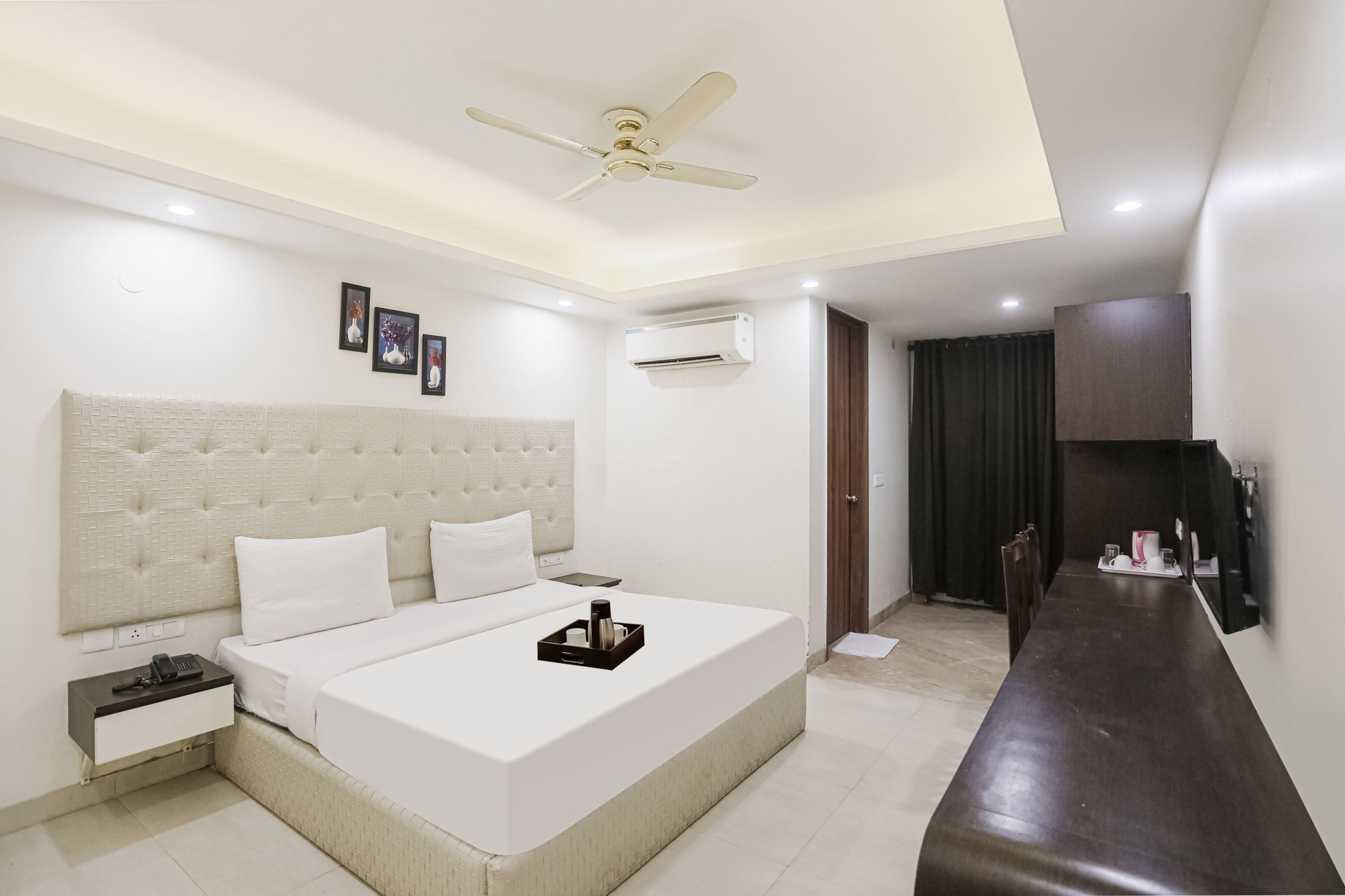 A hotel room with a large bed, a bedside table, an air conditioning unit, a ceiling fan, and a view of the dining area.