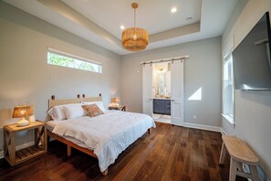 A bedroom with a large bed, bedside lamps, a ceiling light, a window, and a door leading to a bathroom. - Designer Home w/ Garage, Fenced Yard & Patio (Houston)
