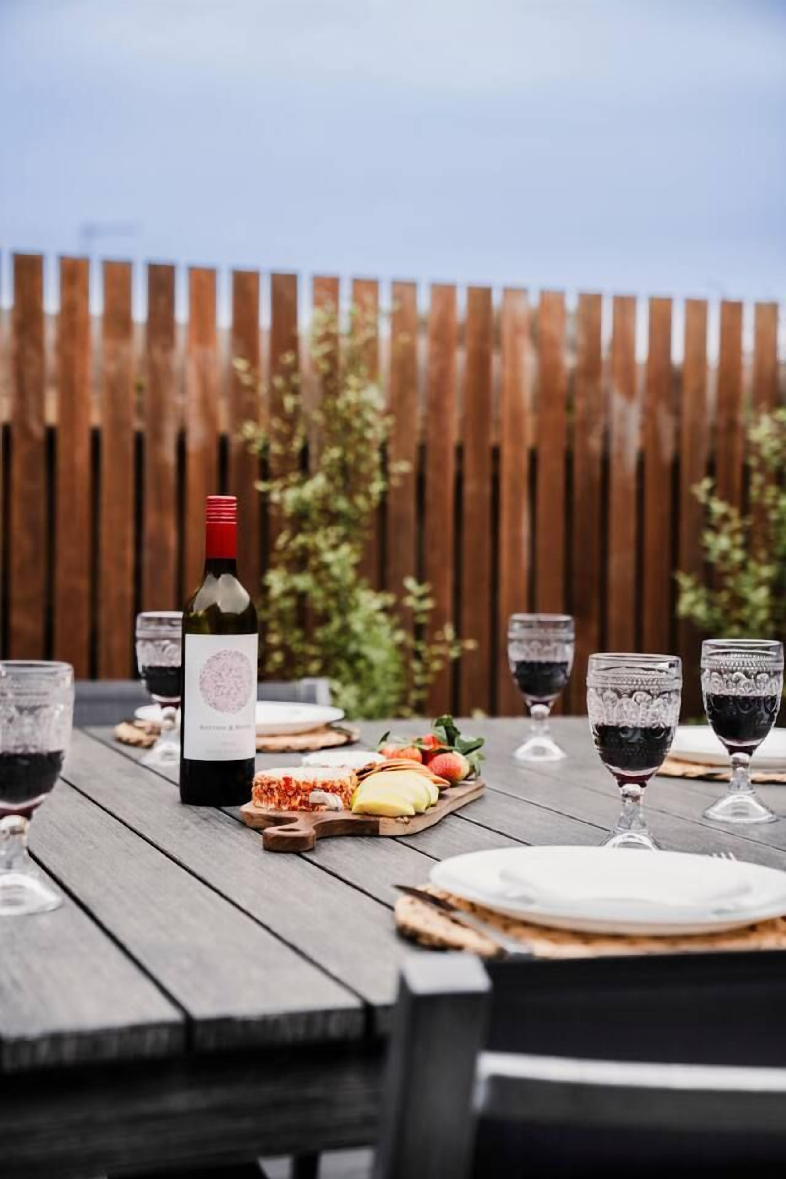 A wooden table set for a meal with a bottle of wine, glasses, and a cheese board.