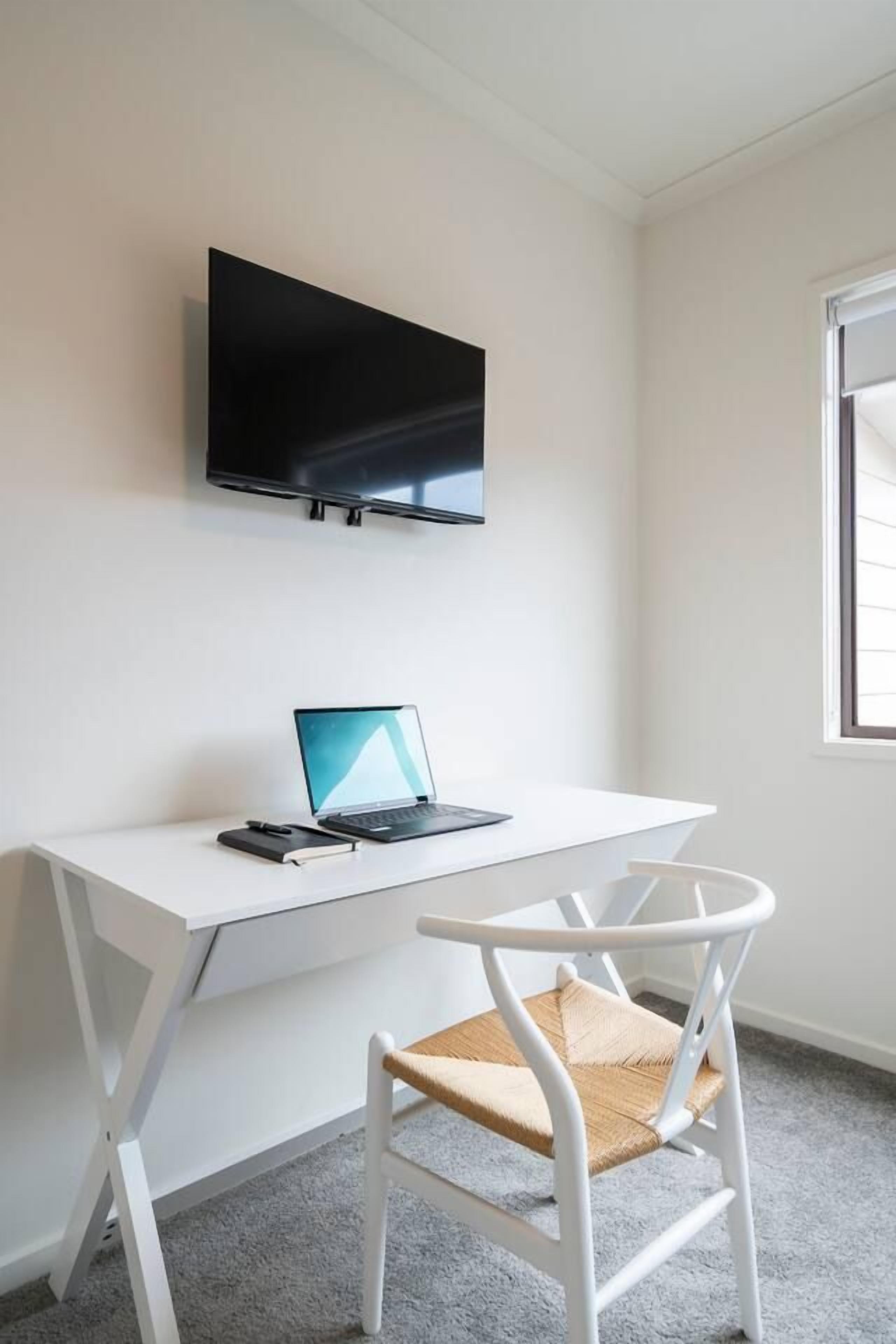 A white desk with a laptop, a flat-screen TV mounted on the wall, and a white chair with a woven seat.