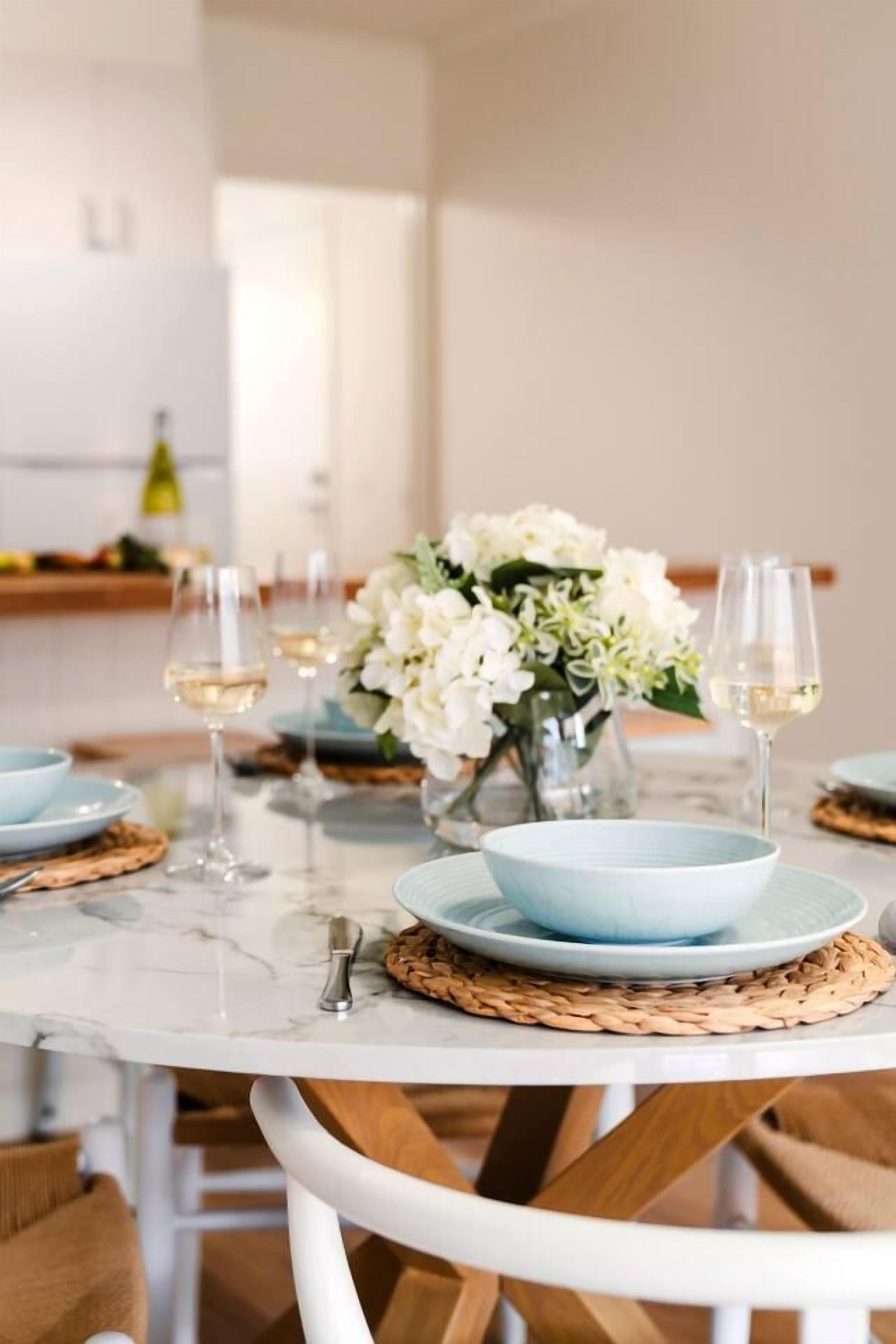 A round dining table with white chairs, set with blue plates, white wine glasses, and a centerpiece of white flowers.