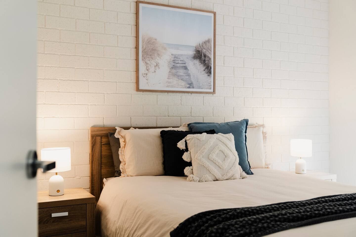 A neatly made bed with a white and blue pillow arrangement, a wooden headboard, and a white brick wall with a framed picture above.