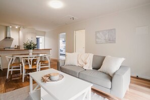 A modern living room with a grey sofa, white coffee table, and a dining area with a white table and chairs. - River Grove Retreat | 100m to River | Pet Friendly (Ocean Grove)