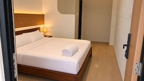 En pænt redt seng med et sammenrullet hvidt håndklæde, en sengetavsalampe og en træsenggavl i et moderne hotelværelse.