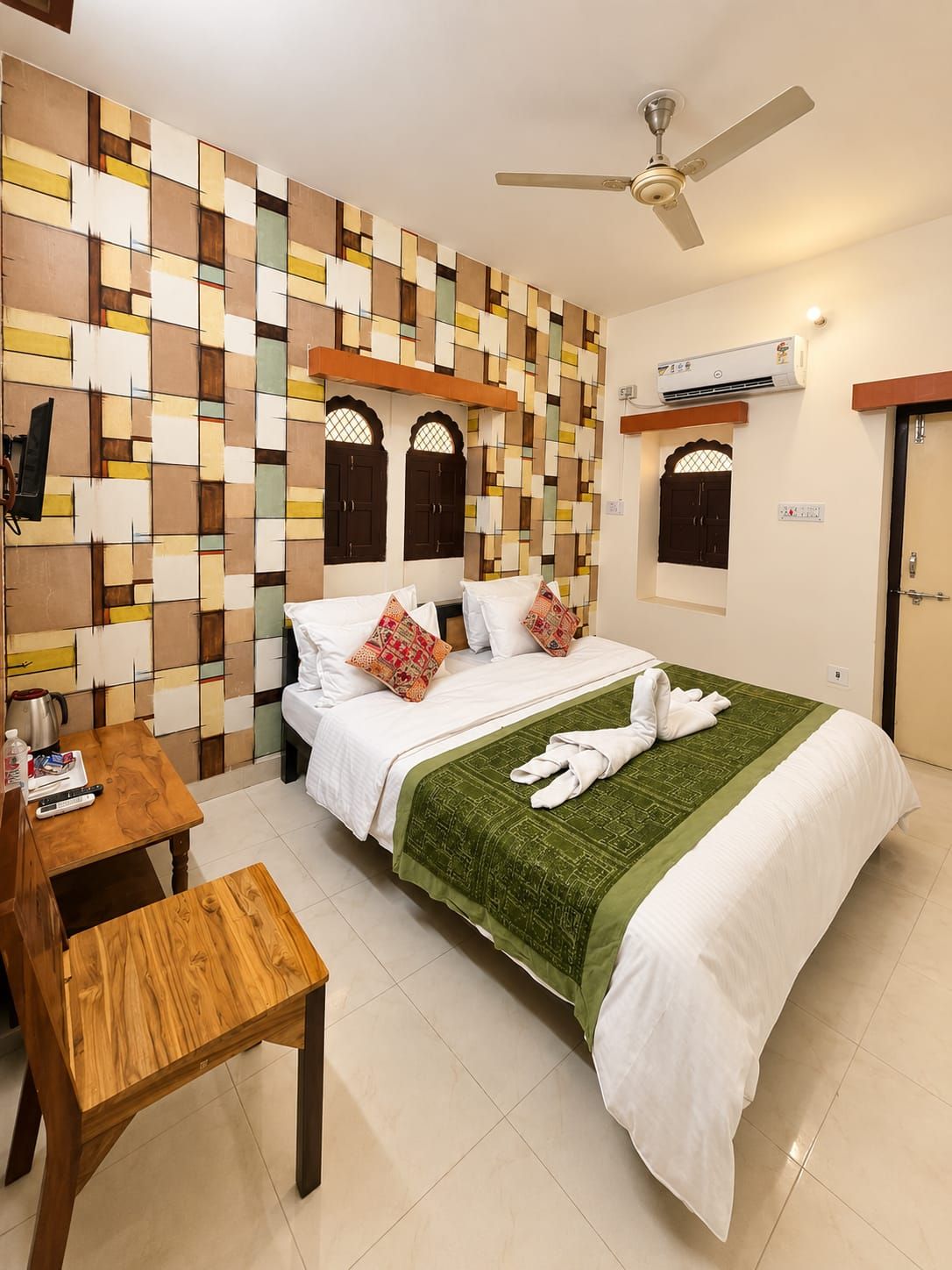 A hotel room with a bed, a wooden table, a ceiling fan, and an air conditioning unit.