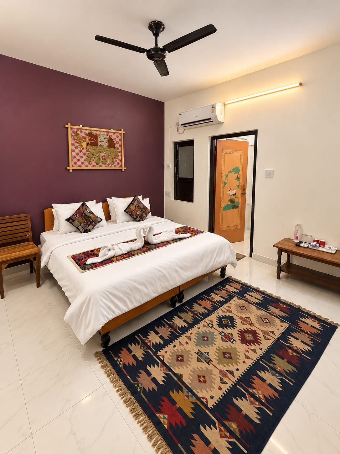 A bedroom with a bed, a ceiling fan, an air conditioning unit, a wooden bench, a small table with items, and a patterned rug.