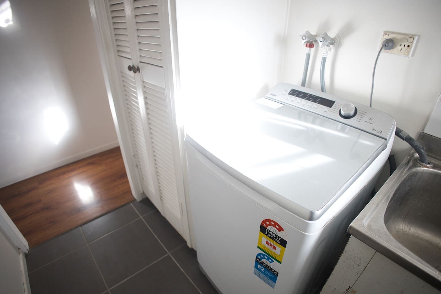 A compact, white washing machine with a digital control panel, situated in a small laundry area with a tiled floor and a wooden door.