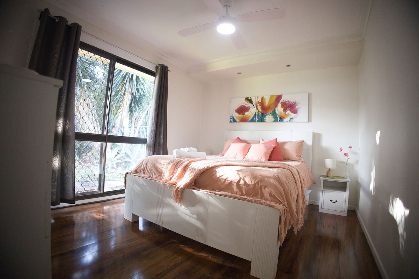 A bedroom with a bed, a nightstand, a ceiling fan, a window with curtains, and a painting on the wall.