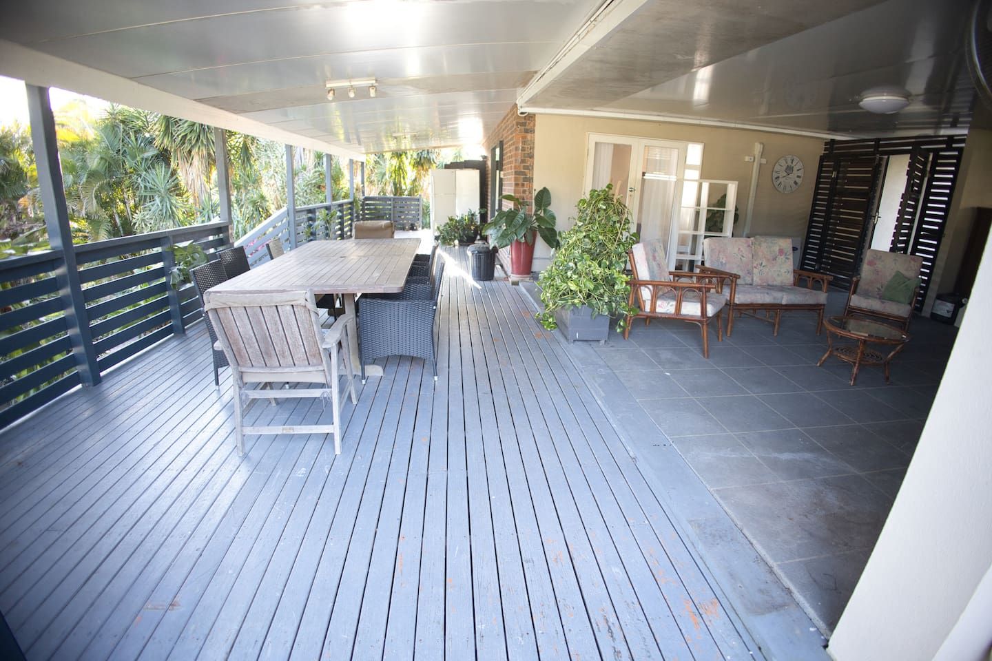 A covered outdoor patio with a wooden table and chairs, surrounded by potted plants and a view of the outdoors.