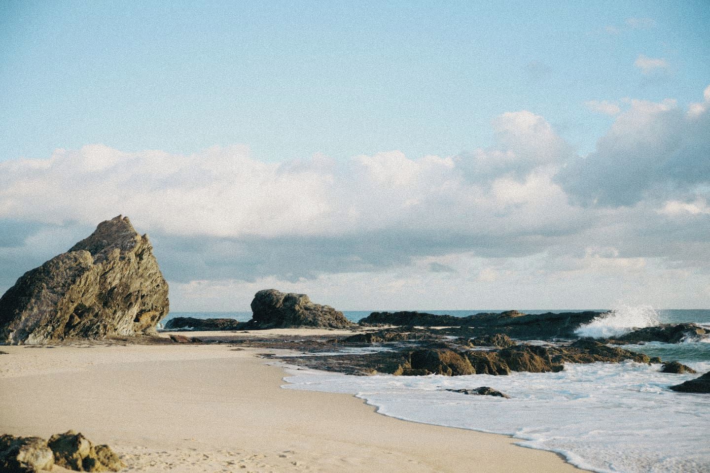 A beach with a large rock formation, sandy shore, and waves.