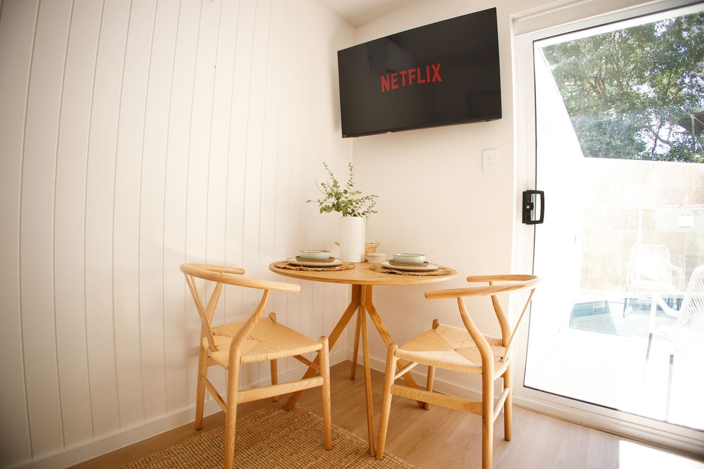 A dining area with a table and chairs, a wall-mounted television, and a window with a view of trees.