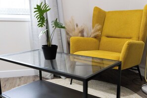 Een moderne woonruimte met een glazen salontafel, een gele fauteuil en een potplant.