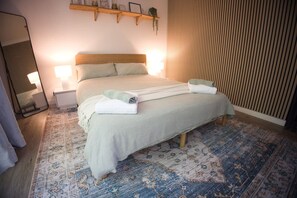 Una cama bien hecha con cabecera, dos lámparas de noche, una mesita de noche y una alfombra estampada.