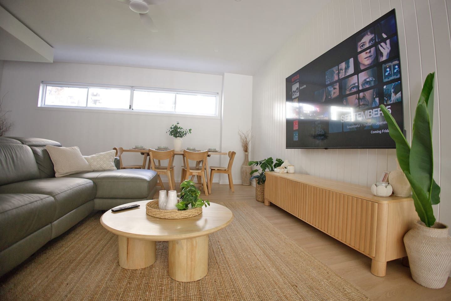 A modern living room with a sectional sofa, a wooden coffee table, a TV mounted on the wall, and a dining area with wooden chairs and a table.