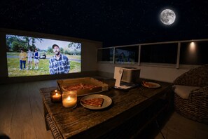 A cozy room with a large screen displaying a movie, a pizza box, a candle, and a slice of pizza on a wooden table.