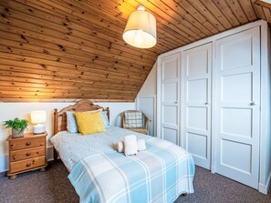Ett sovrum med ett träpanelat tak, en säng med ett blå- och vitrutigt överkast, ett träbord som sängbord med en lampa och en vit garderob.