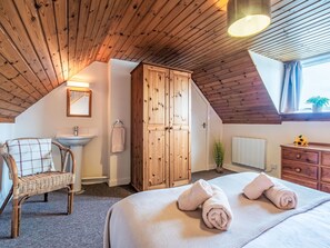 A cozy bedroom with a sloped ceiling, wooden wardrobe, a bedside table, a chair, and a window with curtains. - Bunloit Farmhouse by Interhome (Drumnadrochit)