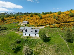 Een landelijk landschap met een wit huis, stenen gebouwen en bloeiende gele struiken.