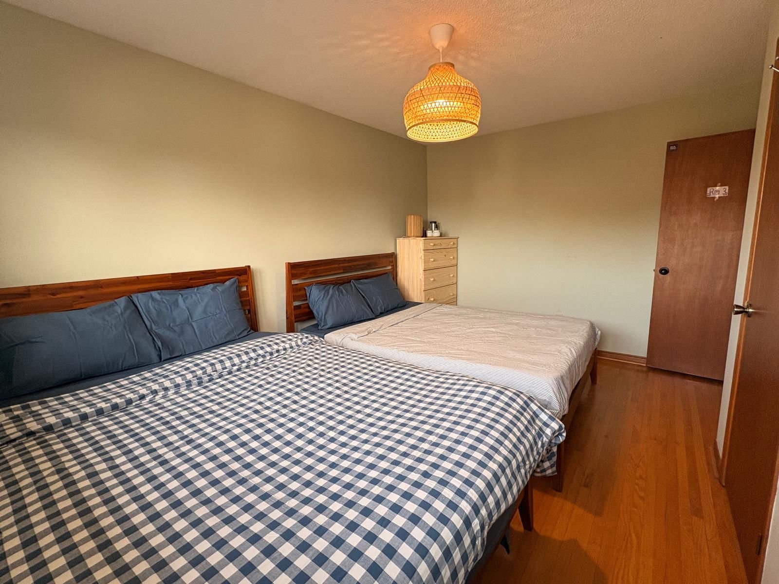 A room with two beds, a wooden dresser, a wardrobe, and a hanging light fixture.