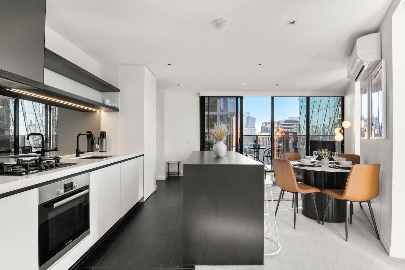 A modern kitchen with a central island, dining area with chairs, and large windows offering city views.